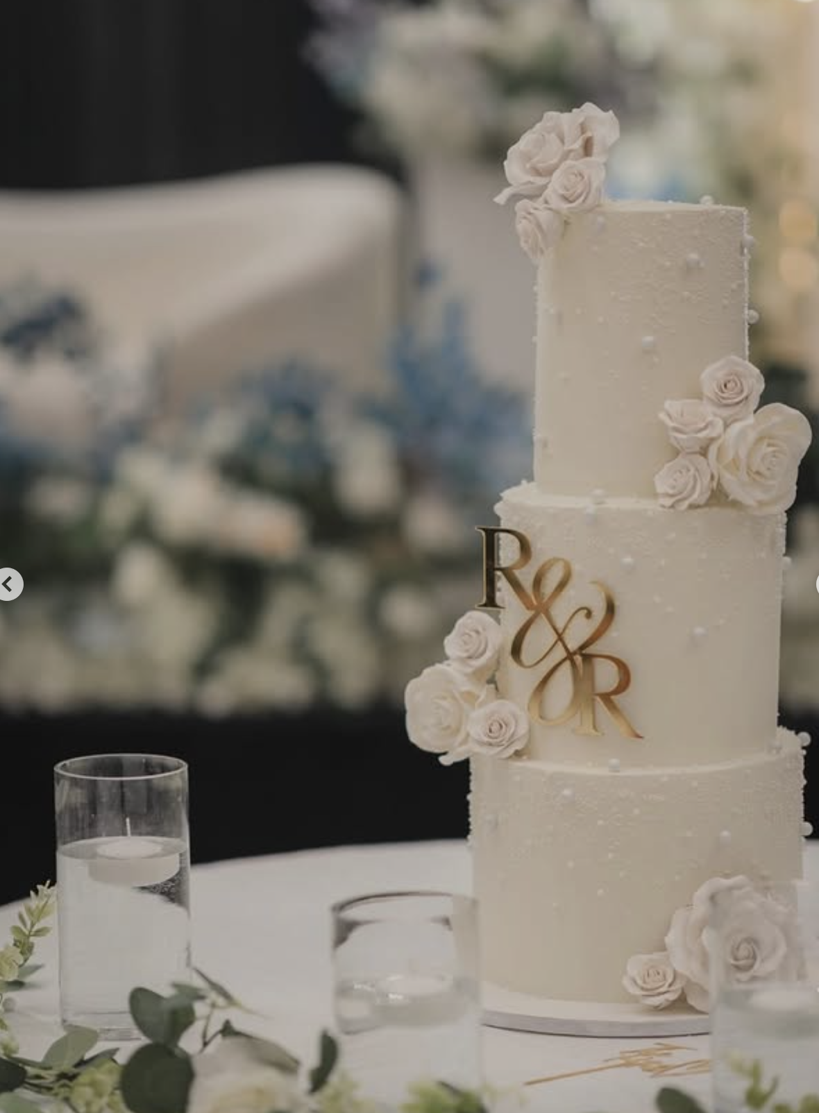 Three-tier white buttercream wedding cake with pearl detail, white sugar roses, and gold R&R monogram topper